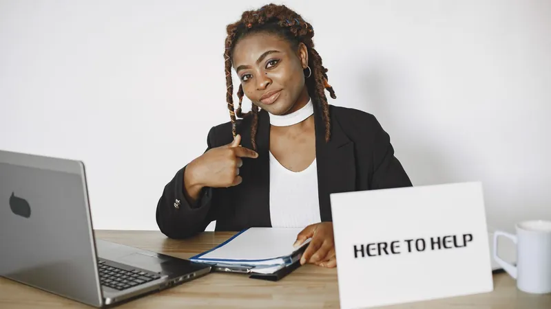 A woman in a black blazer sits at a desk with a laptop and a folder, pointing to herself beside a card that reads “HERE TO HELP”.