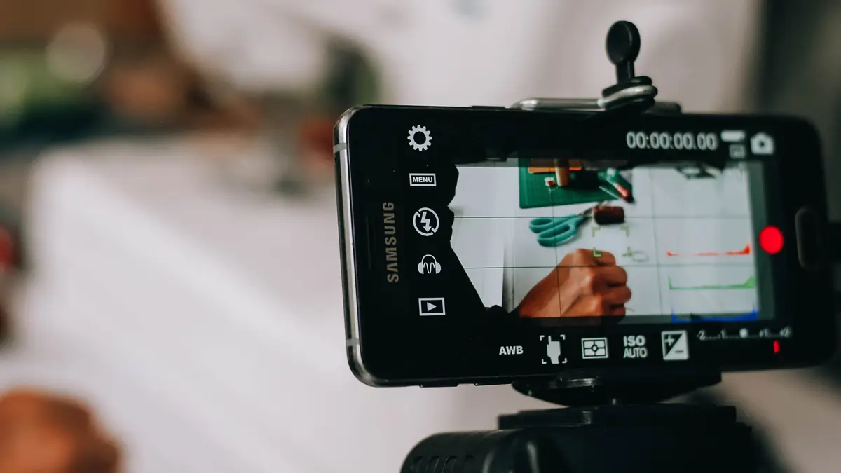 A smartphone held sideways in a tripod clamp, its camera app framing a hand with a pen over a sheet of paper beside green scissors and a cutting mat, the timer at zero and the red record button ready