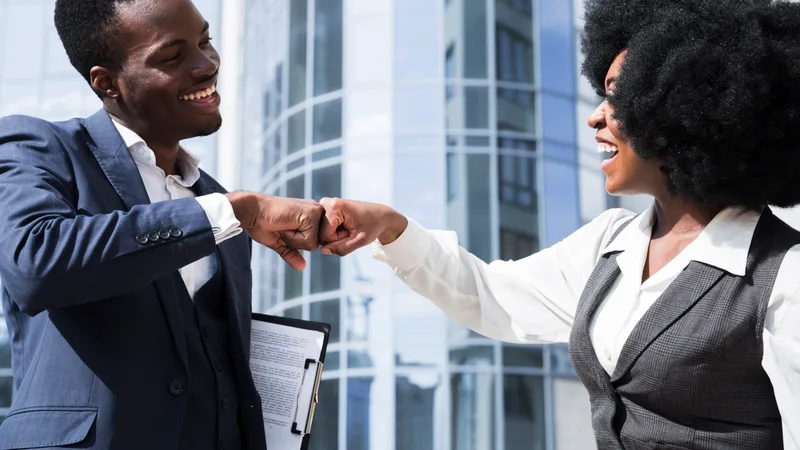 A man in a navy suit and a woman in a grey waistcoat fist-bump outside a glass-fronted office building, both smiling, she holding a clipboard in her other hand.