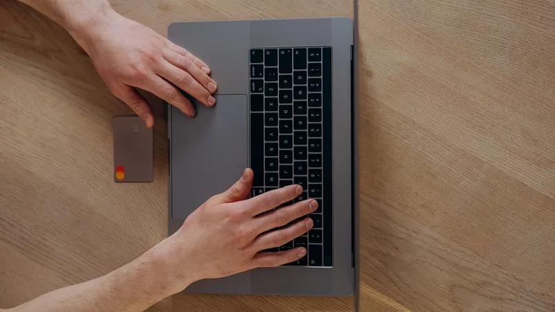 Overhead view of two hands on a laptop’s trackpad and keyboard, a grey bank card lying on the pale wooden desk beside the machine.