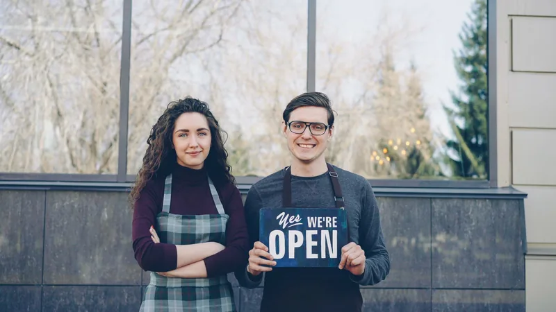 Two shop owners in aprons standing outside their premises on a grey day, one of them holding up a small painted board reading “Yes We’re Open”.