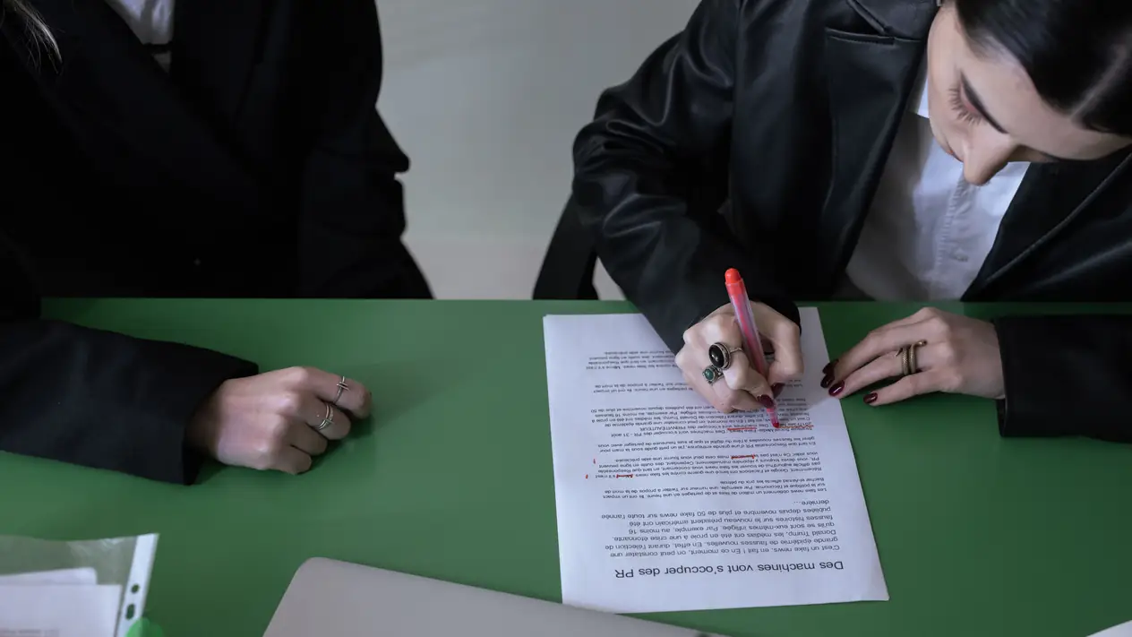 Seen from above, a woman in a black leather jacket marks up a printed page with a red pen at a green table, while a second person in black sits beside her with their hands resting on the table.