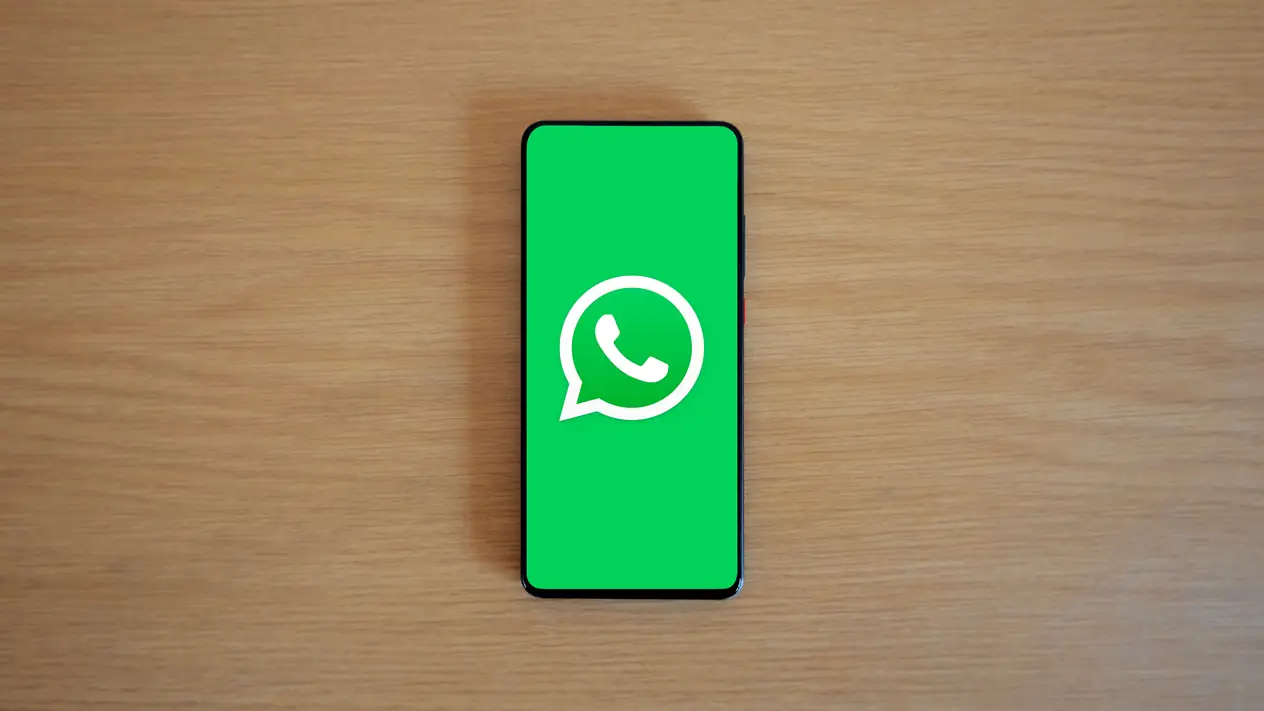 Seen from above, a black smartphone lies on a light wooden desk, its screen filled with the white WhatsApp logo on a green background.