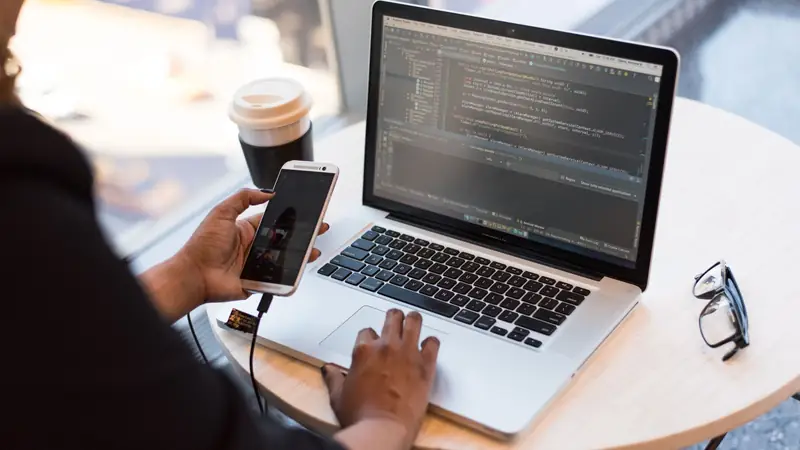 A developer at a round café table types on a laptop showing a code editor while holding a smartphone connected to it by a cable, a takeaway coffee cup and glasses beside the laptop