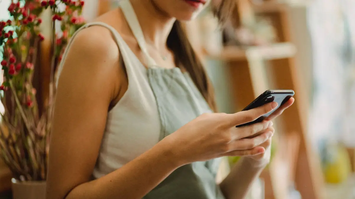 A woman in a sage-green apron over a white top reads a smartphone held in both hands, a vase of red berry stems beside her and the shelves of a small flower shop out of focus behind