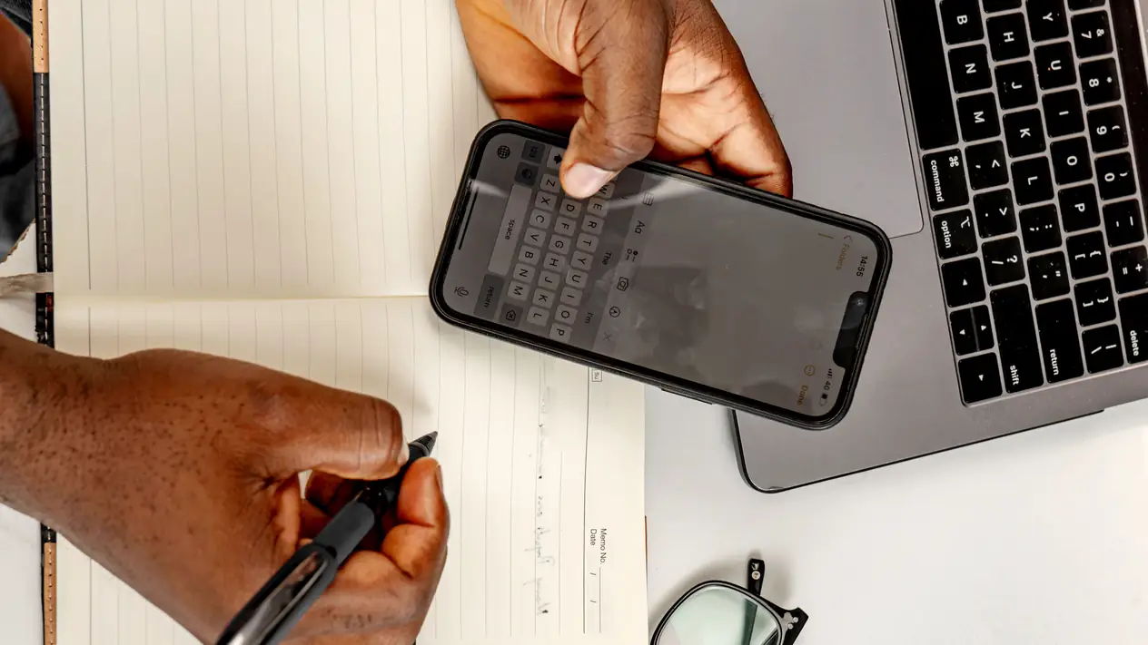 Seen from above, one hand holds a smartphone with its keyboard open while the other holds a pen over a lined notebook, beside a laptop and a pair of glasses.