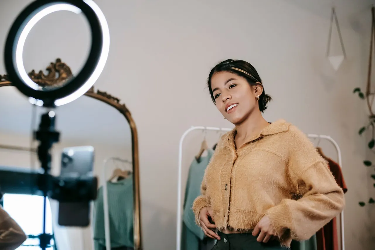 A woman smiling to a phone mounted on a tripod behind a ring light, filming a clip in front of a clothes rail in a small shop
