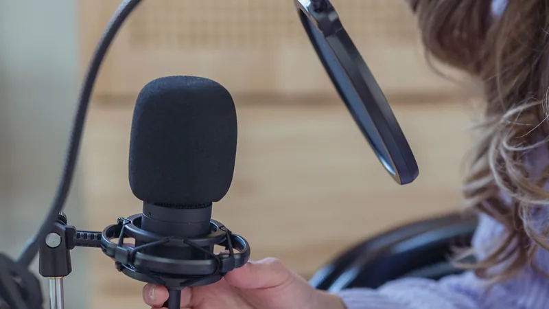 A hand steadying a condenser microphone in a shock mount behind a pop filter, with the speaker seated just out of focus to the right
