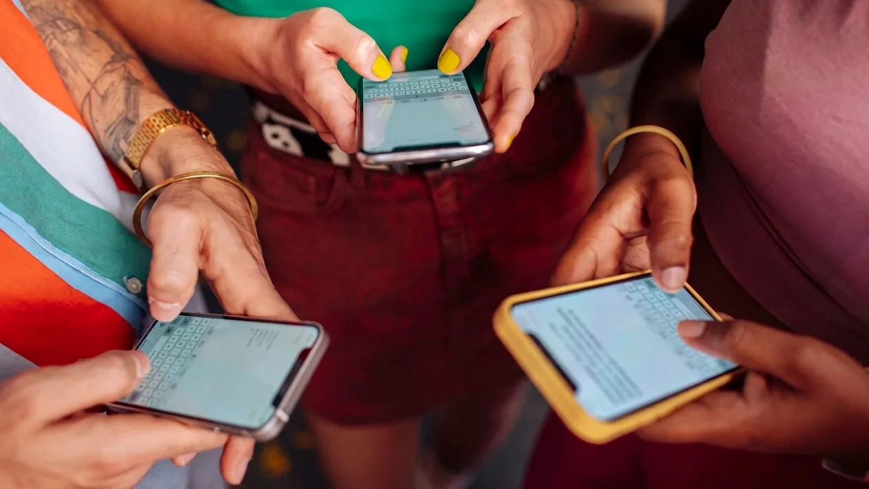 Three people stand close together, each typing a message on their own smartphone, one of the phones in a yellow case.
