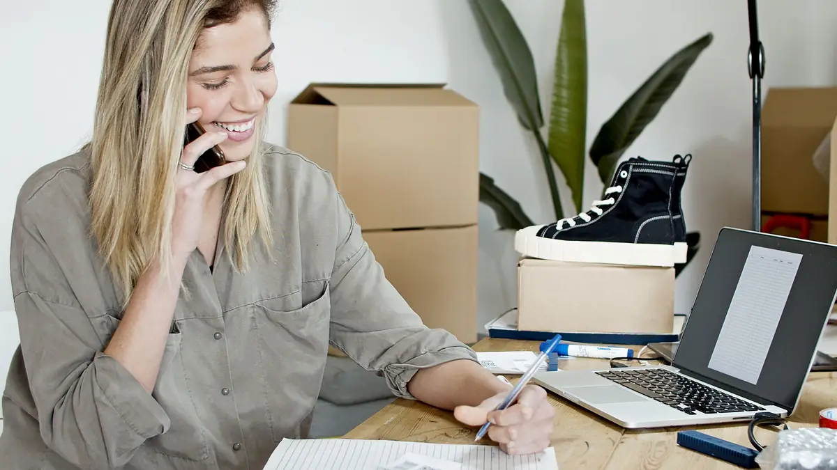 A smiling woman in an olive shirt talks on her phone while writing on an order sheet, a laptop showing a blank form beside her and a black sneaker on a shipping box behind