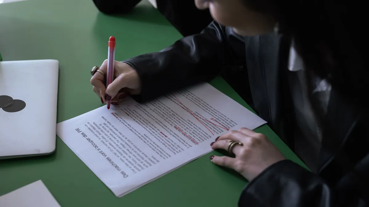 A person in a black leather jacket strikes through a paragraph of a printed article in red pen at a green table, a closed laptop beside the page