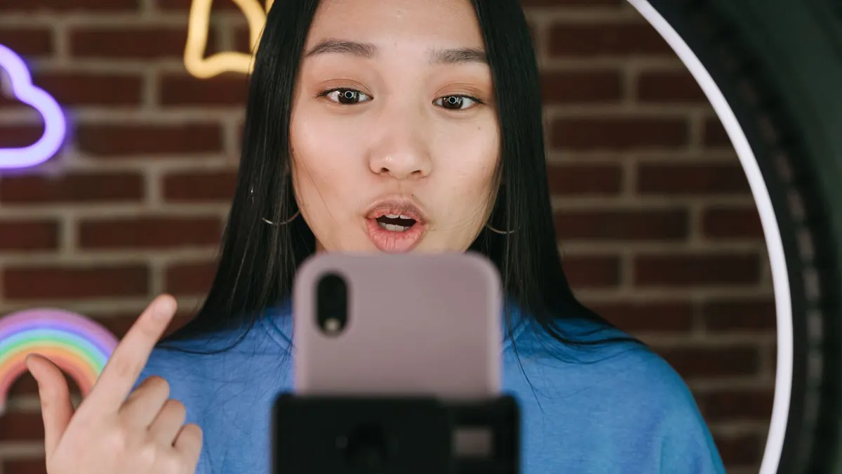A woman in a blue sweatshirt talks into a phone mounted on a stand inside a ring light, mouth open mid-word and one finger raised, with neon cloud and rainbow signs on the brick wall behind her