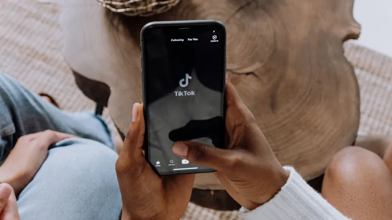 Seen from above, a hand in a white knit sleeve holds a black smartphone showing the TikTok launch screen, thumb over the screen, above a round wooden table and a woven rug