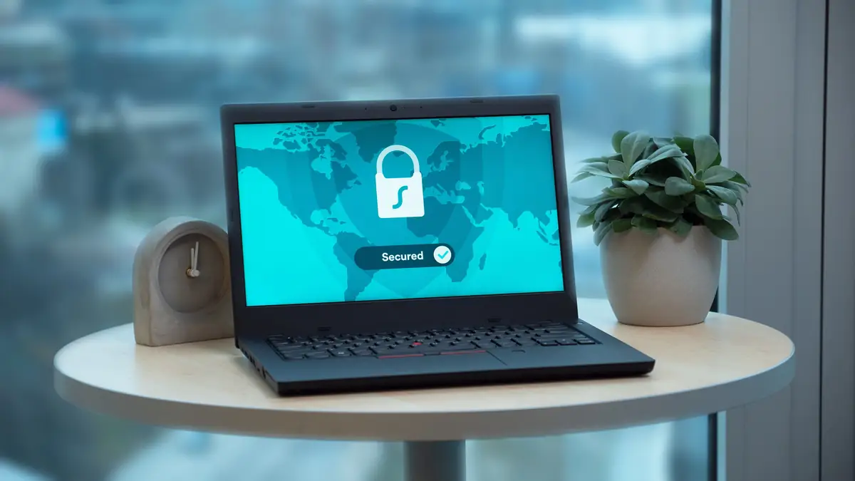 A laptop on a small round table by a window shows a large white padlock and a “Secured” badge over a world map, between a stone desk clock and a potted succulent