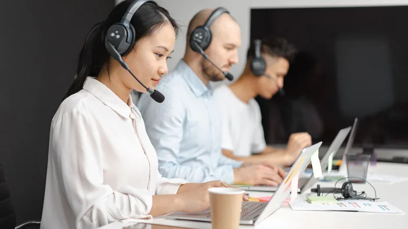Three customer service agents wearing headsets work side by side at laptops in an office, handling support conversations.