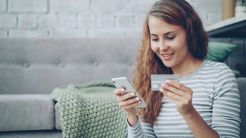 A woman smiles while holding her phone in one hand and a payment card in the other, completing a purchase started through a mobile chat.
