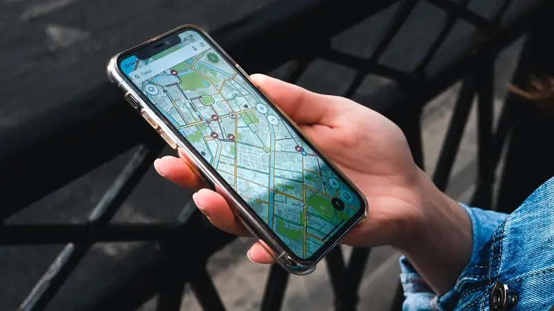 A hand holds a smartphone showing a street map with several pinned locations, beside a dark metal railing.