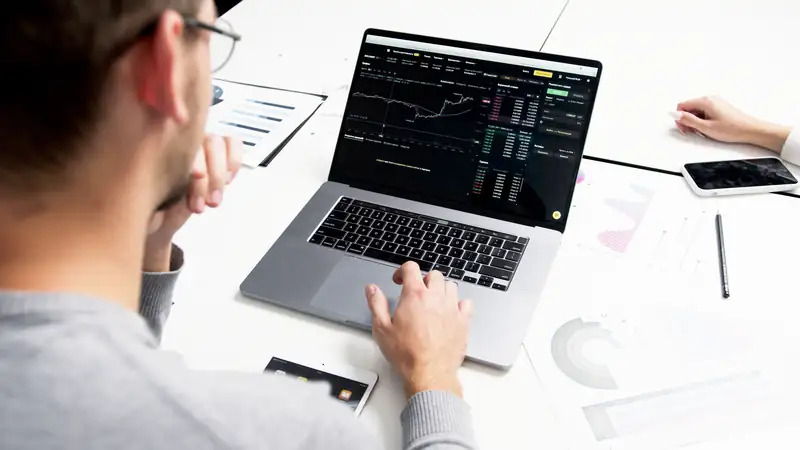 Over a man’s shoulder, a laptop on a white desk shows a dark dashboard with a line chart, next to printed bar and pie charts and a phone.