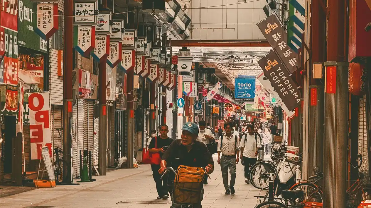 A covered shopping arcade in Osaka lined on both sides with small shops, their hanging signboards and banners stretching into the distance, as a man cycles towards the camera past pedestrians.