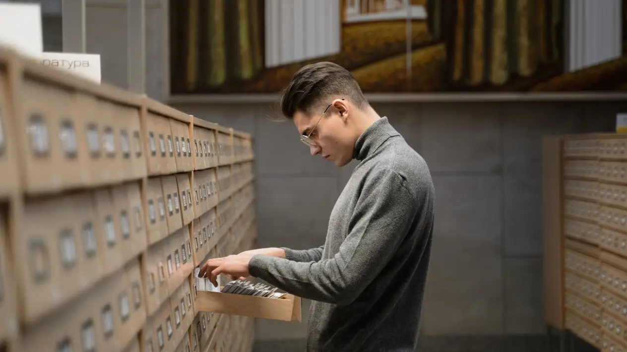 A young man in glasses and a grey roll-neck sweater leans over an open drawer of index cards in a library card catalogue, flicking through the cards, with rows of wooden catalogue drawers on both sides.